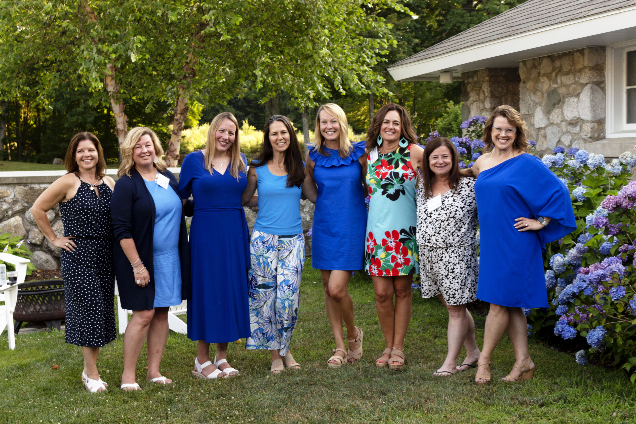 A group of eight women standing on grass in a garden, smiling, with some wearing blue outfits and others in floral patterns. A stone building and hydrangea bushes are in the background. Saint Joseph's College of Maine