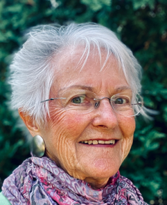 A smiling elderly woman with short white hair, wearing glasses and a patterned scarf, stands in front of a blurred green background, exuding the confidence and grace of someone on the Board of Trustees. Saint Joseph's College of Maine