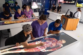Medical students and instructors at Saint Joseph’s College study a 3D digital anatomy table displaying a human body model in a modern classroom equipped with multiple screens and lab equipment, part of the Sciences Department curriculum for the Doctor of Chiropractic Program. Saint Joseph's College of Maine