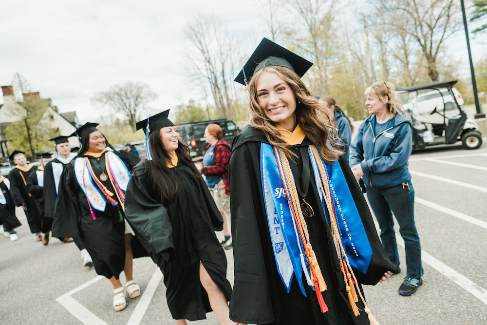 A group of SJC graduates in caps and gowns walk outdoors, led by a smiling woman with long hair wearing a blue sash and several honor cords. The Class of 2024 celebrates amidst trees and parked cars in the background. Saint Joseph's College of Maine