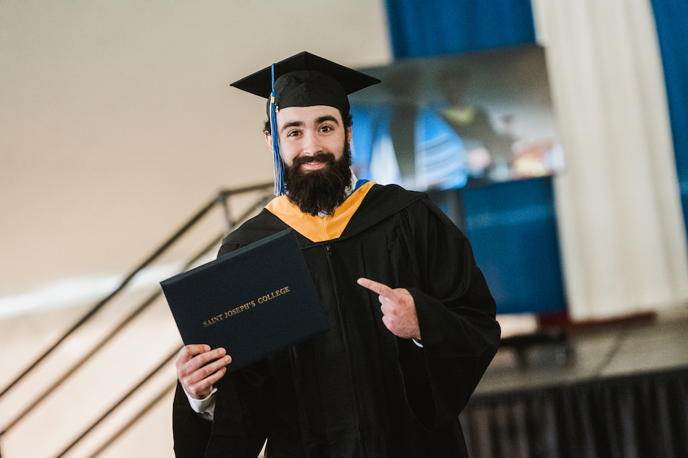 A graduate from the Class of 2024 with a beard and cap points to his diploma while smiling on a stage, celebrating his achievement, with a blurred person in the background. Saint Joseph's College of Maine