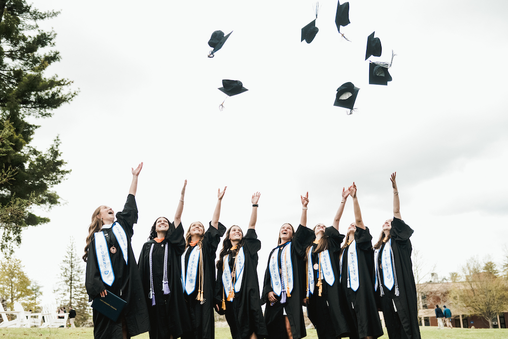 A group of SJC graduates in black gowns and caps toss their caps into the air outside, as the Class of 2024 joyously celebrates their graduation. Saint Joseph's College of Maine