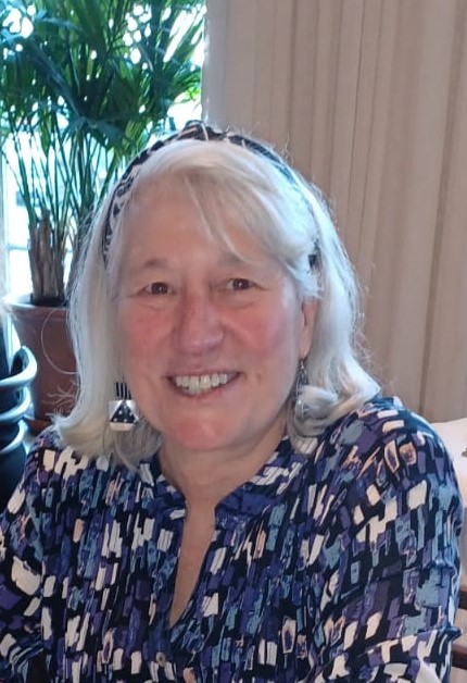 A smiling woman with white hair, wearing a headband and a patterned shirt, sits in front of a potted plant indoors, embodying the approachable demeanor of the President of our Leadership Team. Saint Joseph's College of Maine